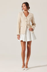 A woman is wearing a cream-colored textured jacket with gold buttons, paired with a white pleated skirt. She has white flat shoes with bows and is standing against a plain light background.